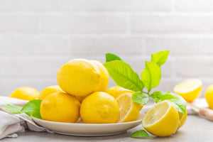 A pile of fresh yellow lemons and green leaves on a white plate against a brick wall.