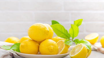 A pile of fresh yellow lemons and green leaves on a white plate against a brick wall.