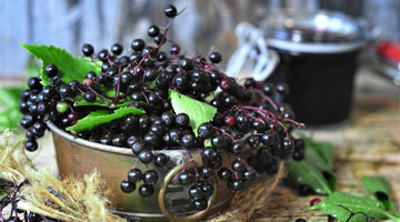 Fresh elderberries with green leaves in a rustic metal bowl on wooden surface with jar in background