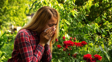 Woman in a red plaid shirt sneezing into a tissue next to red roses in a garden.