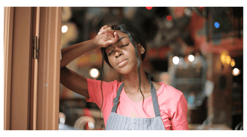 Tired young woman in a pink shirt and gray apron leaning against a doorframe in a cozy restaurant