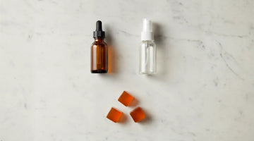 Amber dropper bottle, clear spray bottle, and three orange botanical lozenges on white marble.