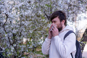 Man blowing nose with a tissue while standing in front of blooming spring trees.