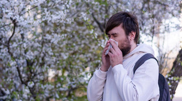 Man blowing nose with a tissue while standing in front of blooming spring trees.