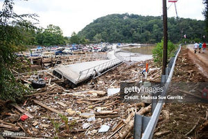 Flood debris and damaged dock near marina with boats, wooded hill, and people walking on a dirt path