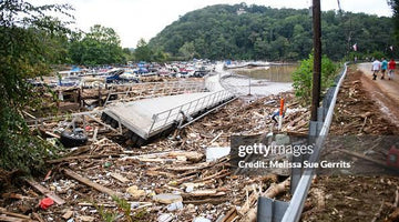 Flood debris and damaged dock near marina with boats, wooded hill, and people walking on a dirt path