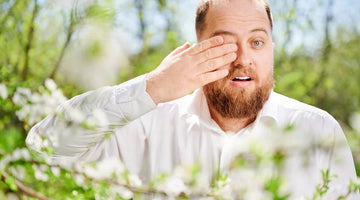 A man with a beard rubbing his eye during allergy season amidst spring blossoms.