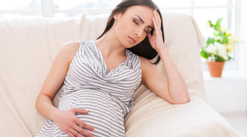 Pregnant woman in a striped dress sitting on a sofa, looking unwell and holding her head.
