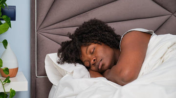 A Black woman sleeping peacefully in a bed with a grey tufted headboard.