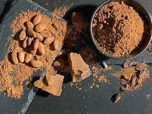 Cocoa beans, cocoa powder in bowl, and broken cocoa chunks on dark slate surface