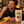 Load image into Gallery viewer, A man in a navy henley shirt is shown eating a piece of chocolate while holding a stand-up pouch of Essential Candy Cacao Orange plant-powered hard candy. The scene is set on a wooden table with fresh oranges, chocolate pieces, and a small bowl, with a warm, cozy background.
