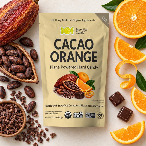 A stand-up pouch of Essential Candy Cacao Orange plant-powered hard candy is centered on a light stone surface, surrounded by cacao beans, a cacao pod, orange slices, orange peels, and chocolate squares. The package is cream-colored with bold black and green text and a tropical cacao-and-orange graphic.