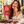 Load image into Gallery viewer, A woman sitting at an outdoor cafe holds up a red bag of Essential Candy Cacao Sea Salt Cayenne hard candy while eating a piece of chocolate candy. The table is decorated with chocolate pieces, red chili peppers, and bowls of ingredients.

