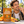 Load image into Gallery viewer, A smiling man outdoors holds a piece of orange cinnamon candy near his mouth next to a large bag of Essential Candy Orange Cinnamon hard candy, with oranges, cinnamon sticks, and candy pieces on a stone table in a sunny urban setting.
