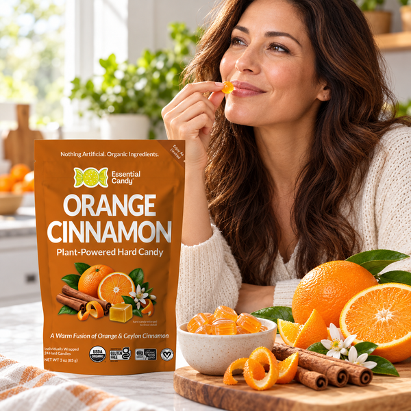 A woman in a cream knit sweater is shown eating an orange cinnamon hard candy while a stand-up pouch of Essential Candy Orange Cinnamon candy sits beside her on a kitchen counter with oranges, cinnamon sticks, and a bowl of candy.
